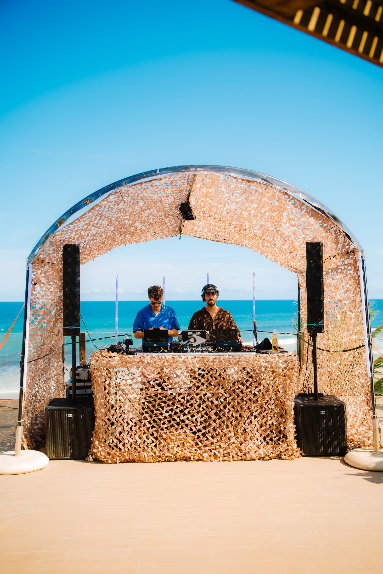 DJs da Drop Music Brasil tocando à beira-mar no Lôro, Salvador, com céu azul e o mar ao fundo